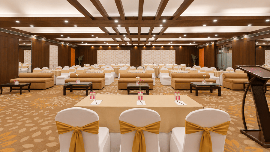 Elegant banquet hall with gold bow-tied chairs, wood ceiling beams, floral carpet, and conference setup.