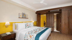 A bright hotel room with a queen bed featuring floral and teal accents, a wooden nightstand with a lamp and phone, a shelf with framed pictures, wooden wardrobes, and an open doorway to a bathroom with yellow walls - Aloha On the Ganges.