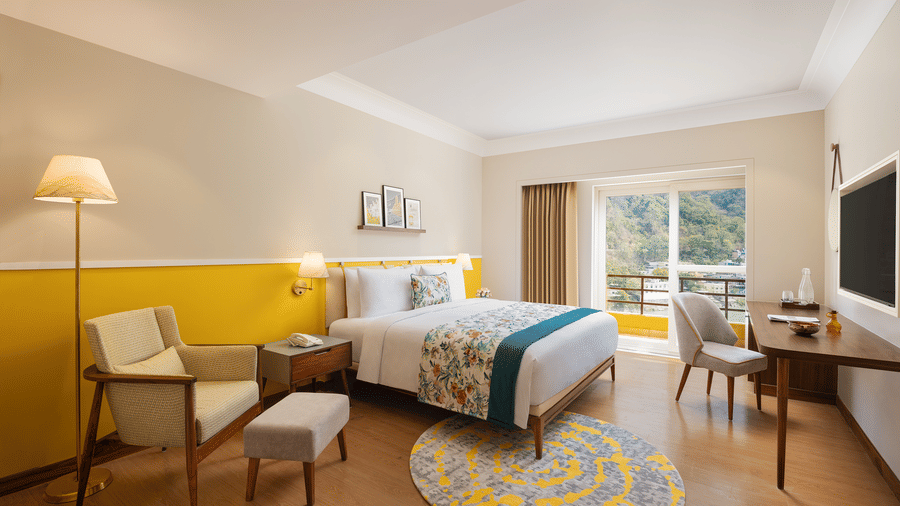 Modern bedroom at Aloha on the Ganges Resort with a large bed, cheerful yellow and beige walls, patterned throw, armchair, desk, and balcony view of green hills.