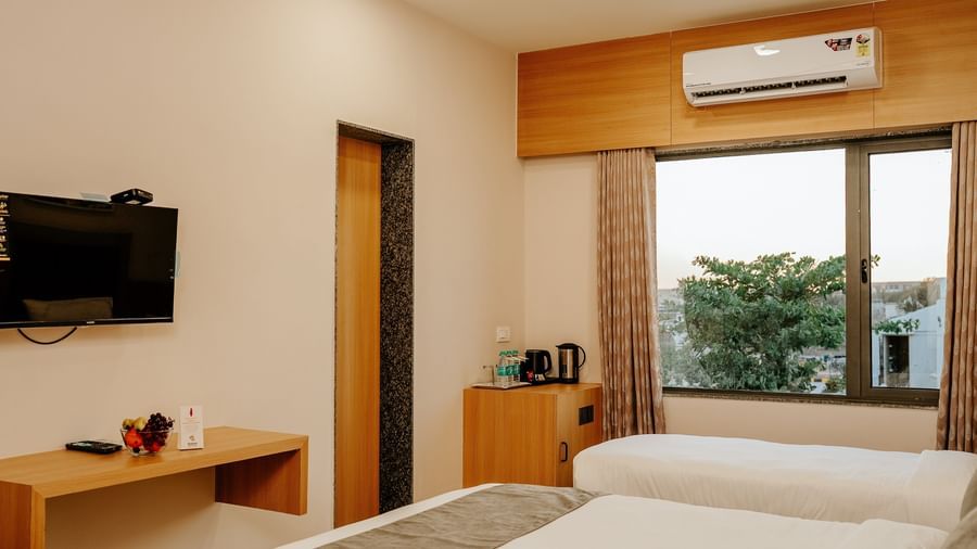 Well-appointed bedroom at Daksh The Grand Gowardhan, Dwarka, showcasing the beds, a wall mounted television, and a window with views.