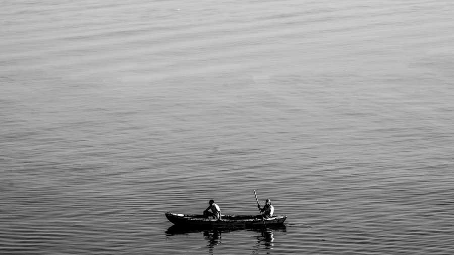 Black and white image of two people rowing a small boat on a vast calm river with gentle ripples visible across the water surface.