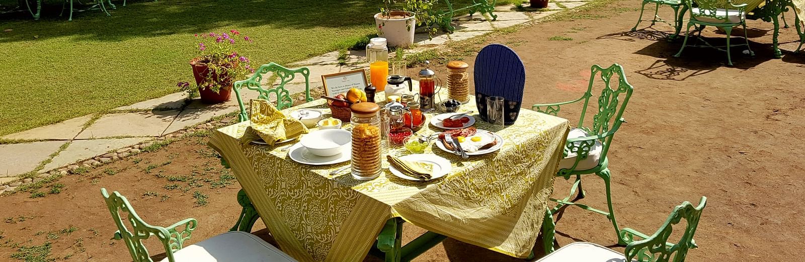An outdoor dining area with tables set for a meal, surrounded by trees and a grassy field, with a person seated in the distance- Shahpura Bagh.