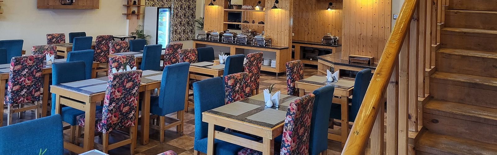 A well-lit, high-ceiling restaurant with wooden interiors, blue cushioned chairs, and an open staircase at Dagshai Manor Kasauli - Inde Hotels.