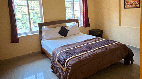 Guest room at Kollanoor Guest House with a double bed, a quilted spread, a ceiling fan, a round window, maroon curtains, and a framed horses painting.