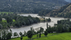 A shot of serene valley with mountains on either side with lush green grass, winding alongside a sparkling river under a clear blue sky.