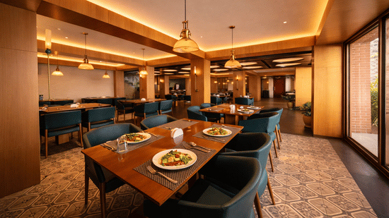 A restaurant dining area with wooden tables, armchairs, and warm cove lighting along the ceiling | Pride Premier Darjeeling