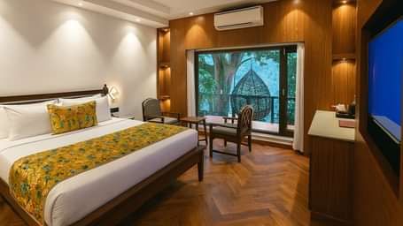 Premium Ganges View Room at Summit by the Ganges Beach Resort and Spa, Rishikesh featuring a double size bed, a sitting area with chairs and centre table, a cabinet table, a flat TV screen, an AC, and a balcony attached which has a chair placed.