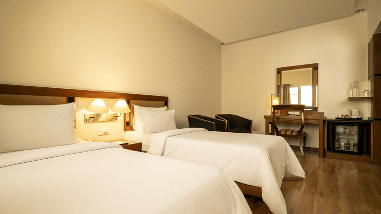 A warmly lit hotel room presents two single beds with white linens, a wooden desk with a mirror, and a mini-fridge at The Residency Karur.