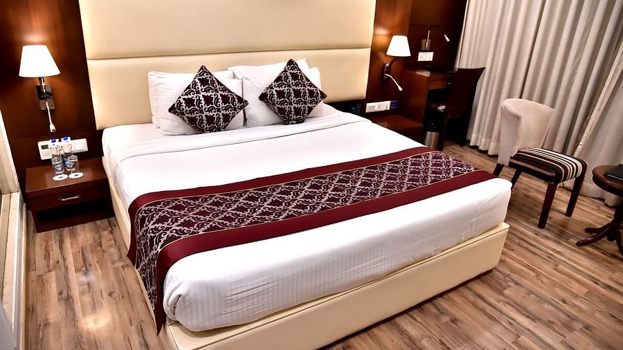 Room at Clarks Inn Suites, Near Gwalior DD Mall featuring a large double bed with cushions and runner, side tables with lamps, work desk, two chairs, round table, and wooden flooring.