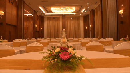 Polo Hotel, Agartala banquet hall view down a white-linen table topped with pink gerbera centerpiece and glass bottle, rows of chairs and chandelier.