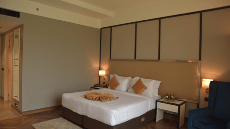 Guest room with king bed, beige headboard, folded towel fan, wing chair and herringbone floor at Polo Hotel, Agartala.