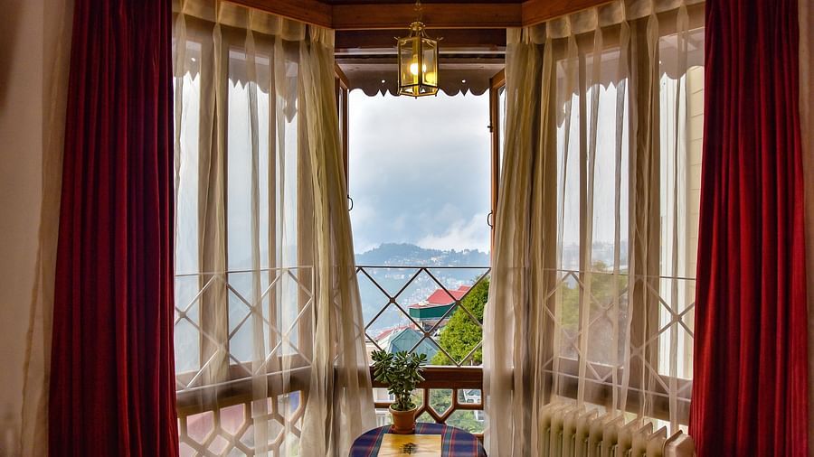 The double-layered red and transparent curtains on the glass window at Cedar Inn in Darjeeling offering a view of hilly landscapes.