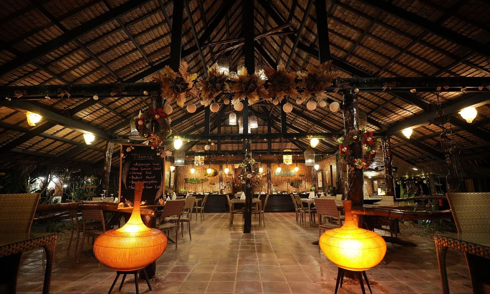 Interior of Flower Island Resort restaurant with wooden furniture and yellow pendant lights under a thatched roof.
