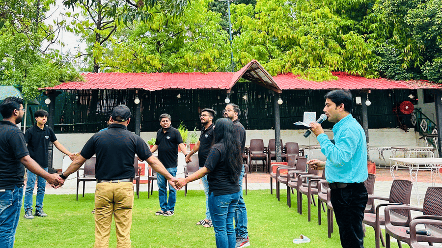 A group of people stand in a circle holding hands on a grassy lawn area surrounded by trees at Amantra Shilpi Resort, Udaipur.