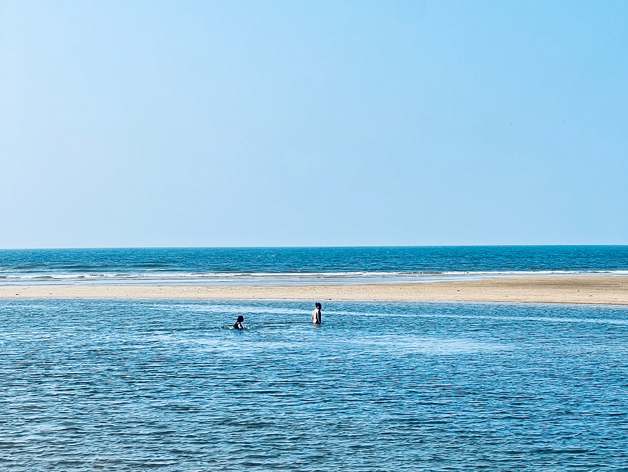 A beach scene featuring two people wading in the water, with the sun shining and waves lapping at the shore.