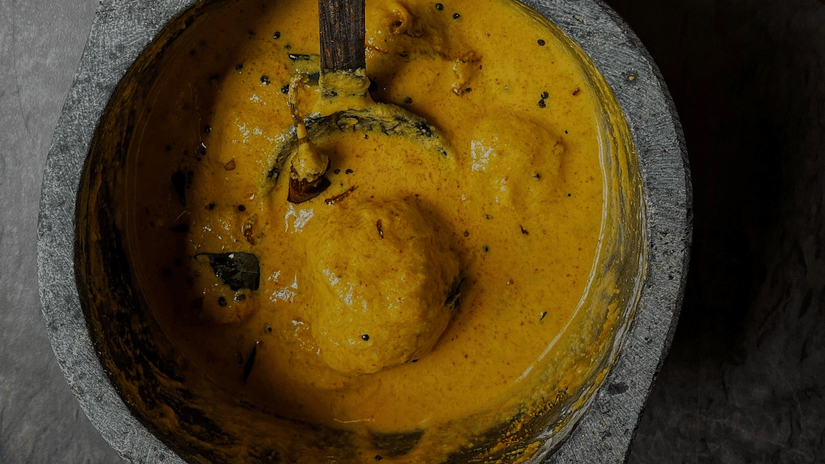 Close-up of traditional Rajasthani gatte ki sabji, featuring gram flour dumplings simmered in a rich, creamy yellow yoghurt-based curry, garnished with spices and served in a rustic bowl.