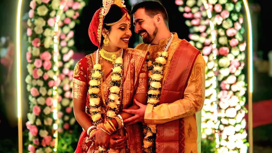 An Indian bride and groom stand together in traditional wedding attire under a beautifully decorated archway, surrounded by floral arrangements