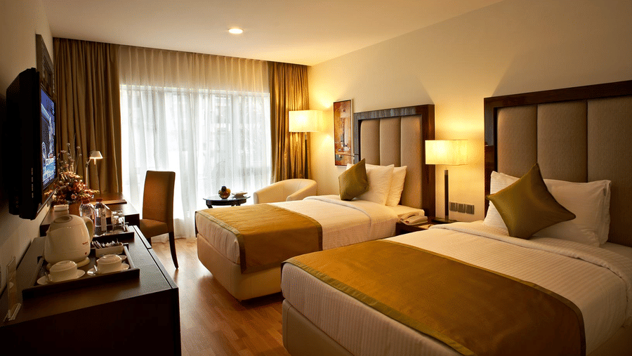 The Executive Twin bedroom featuring twin bed, bedside table with lamps, flat TV screen, a cabinet with tea and coffee maker and a work desk and chair at Adarsh Hamilton.