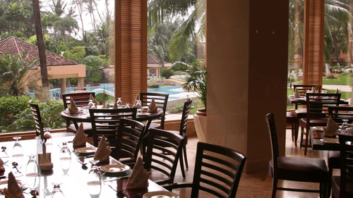 A dining area in one of the restaurants in Madh Island with wooden chairs and tables overlooking a pool area framed by palm trees.