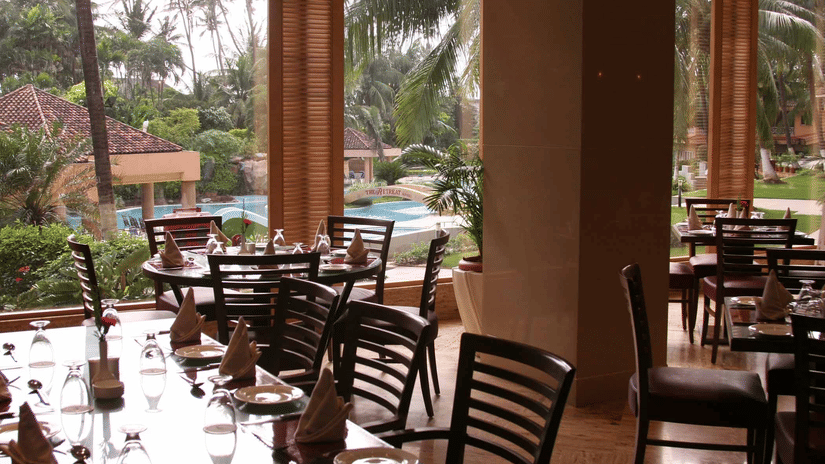 A restaurant dining area with wooden chairs and tables overlooking  a pool area framed by palm trees.