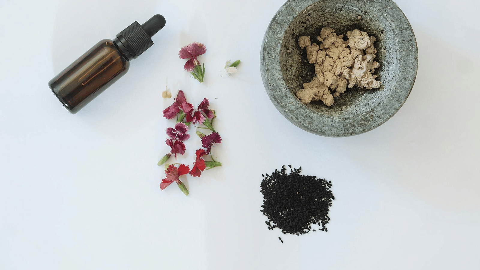 A glass vial, flowers, herbs and a mortar aesthetically placed on a white surface.