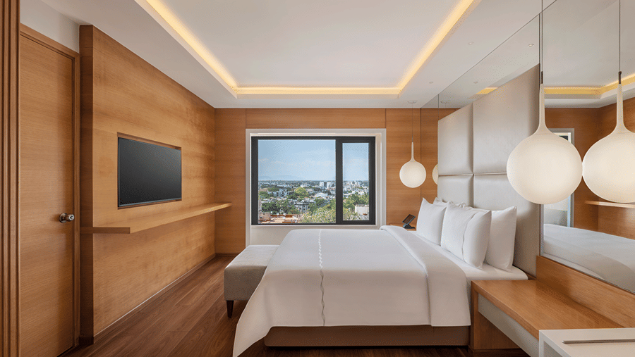 Suite living area with a grey sofa and armchair, round dining table with three chairs, work desk, coffee station and a large window overlooking the city.