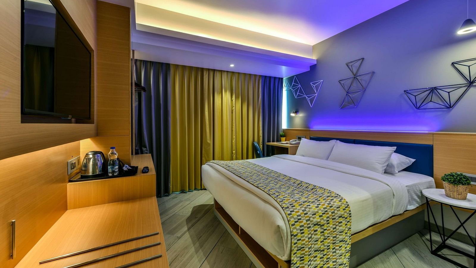 A spacious room with a large bed, a wall-mounted television, a desk positioned below it, and windows with curtains drawn, illuminated by cool lighting at Indie Stays, BKC Kalina.