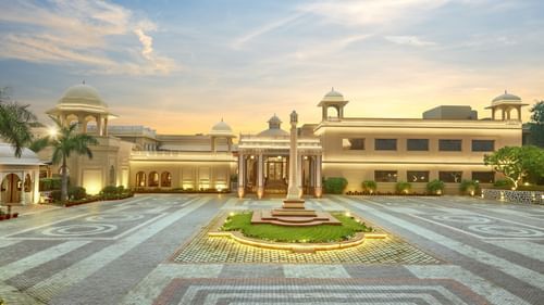 Manesar - Front view of a grand hotel building at dusk, featuring a symmetrical design with a central fountain, landscaped gardens, and ornate light posts leading to the main entrance, all under a soft, glowing sky.