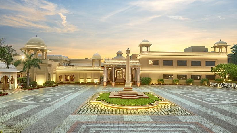 Manesar - Front view of a grand hotel building at dusk, featuring a symmetrical design with a central fountain, landscaped gardens, and ornate light posts leading to the main entrance, all under a soft, glowing sky.
