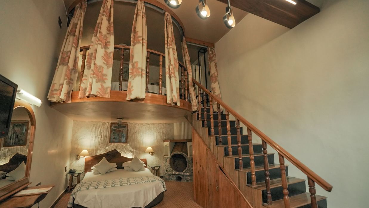 A bedroom with a bed, a rug, and warm lighting, interior setting with wooden elements and stairs - The Den Corbett Resort & Spa, Kumeria.