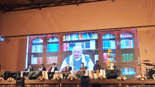 A renowned public figure giving speech on a large projector on the stage as the rest of the members are sitting on the chairs at Uday Palace Convention Centre