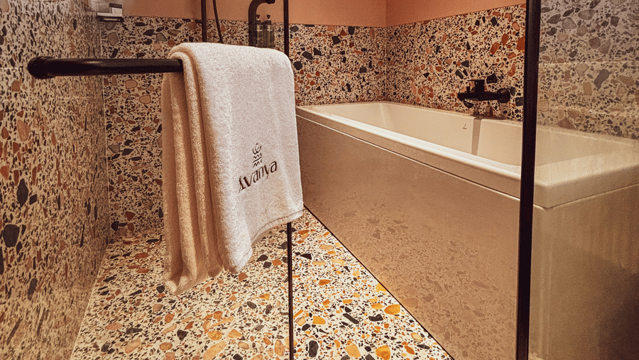 A white bathtub is positioned against a speckled wall and floor behind a glass shower door at Avanya Resorts N Retreat.