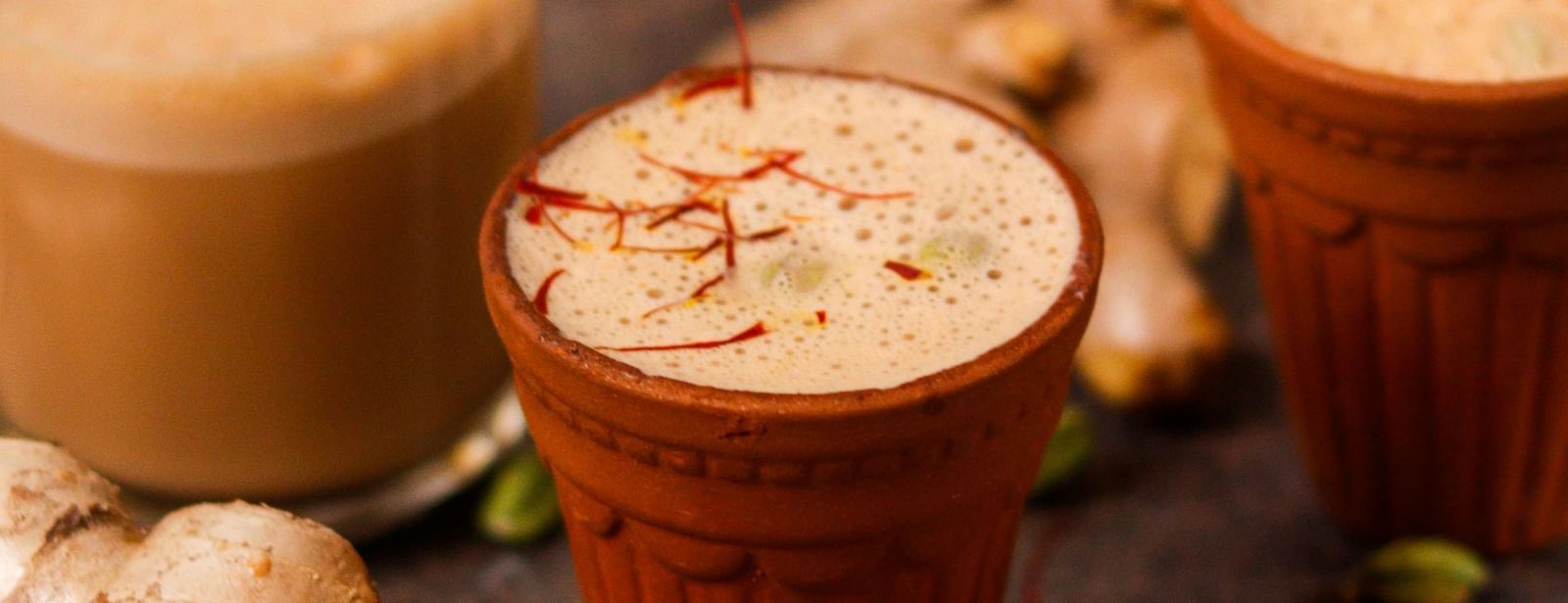 Clay cups filled with tea placed on a dark surface with cardamom, ginger, and a grater scattered around.