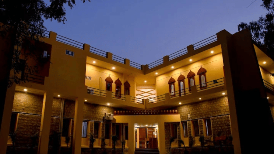 The courtyard and 2-storey exterior of Estherea Bagh, Ranthambore, at dusk, showing stone architecture, lit windows, and a checkered floor pattern under a clear sky.