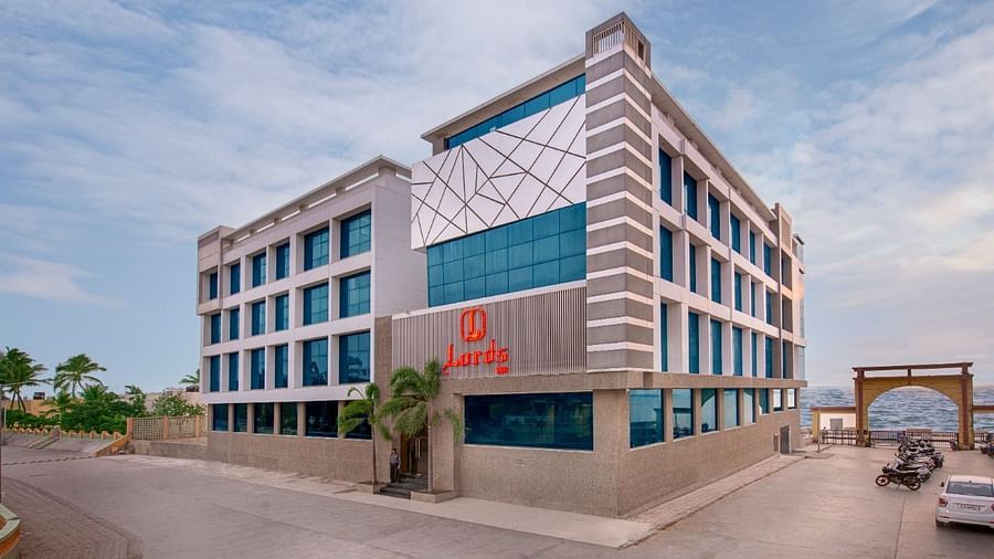 A light-colored hotel with a distinctive architectural style is located near the coast, with gentle waves lapping the shore under a clear sky.