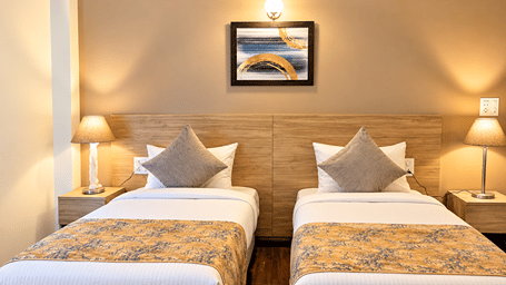 Front view of twin beds in the Premium Studio at Starlit Suites, Shirdi, with grey cushions, patterned runners, bedside tables, lamps and framed artwork.