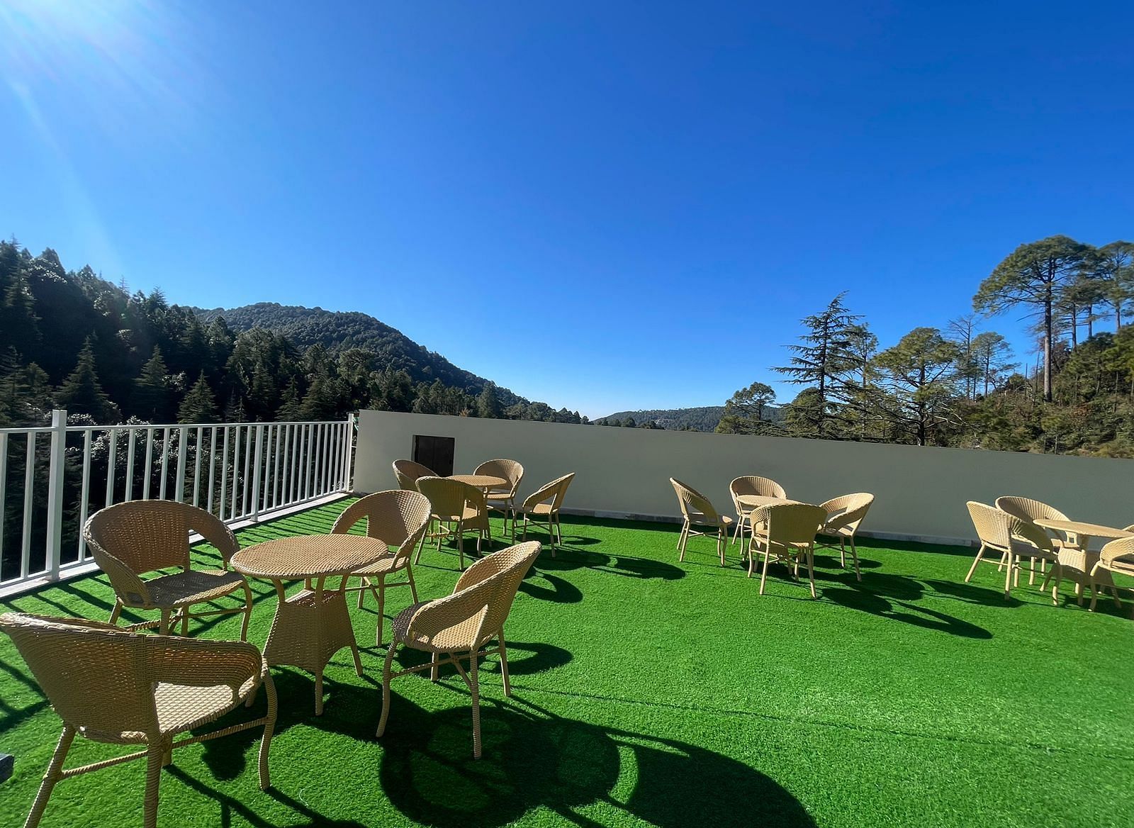 Open terrace area with chairs around tables with a background of trees at DLS Ark Holiday Inn, Banikhet during a sunny day.