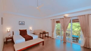 A room featuring a plush double bed, wooden furniture, a spacious seating area, and large glass doors opening to a private balcony with scenic views. - Abad Brookside Lakkidi, Wayanad