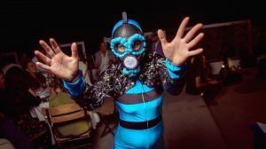 A person wearing a blue outfit and a gas mask stands with their hands raised in a dark environment.