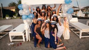 A group of women in blue and white attire, some wearing hats, pose outdoors under a decorated canopy with balloons and white lounge chairs.