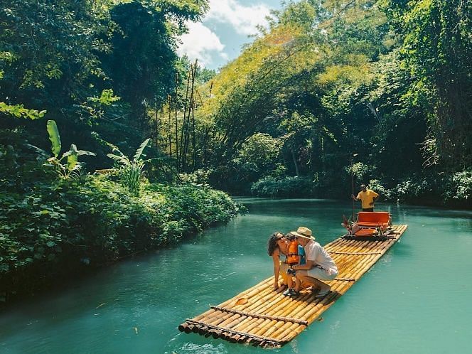 A serene river with clear water flows through a dense tropical jungle, with people on a bamboo raft.