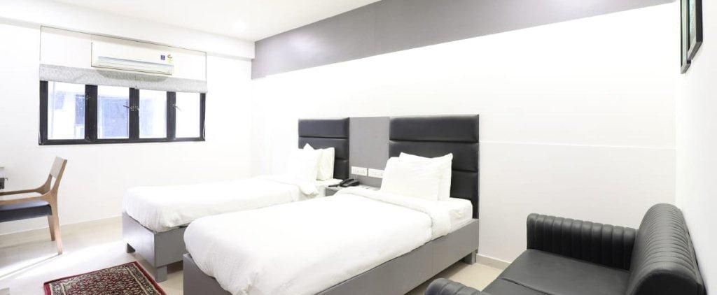 Corner view of the Premium Tin Room at Pravasa Stay, Ahmedabad (Alba Premier), showcasing twin beds along wit a patterned rug in front of it and a black leather couch kept beside it.