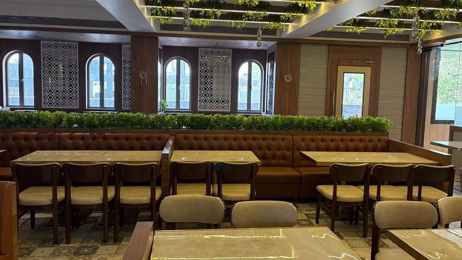 A brightly lit restaurant area with wooden tables and chairs, featuring arched windows and some greenery - Melior Inn