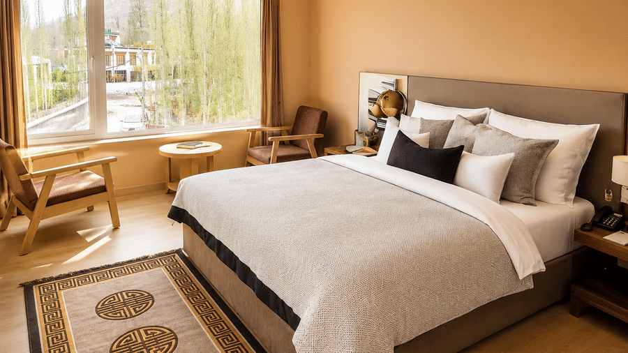 Hotel room with a bed dressed in white linen, a wooden headboard, a patterned rug and a large window overlooking mountain views close by.