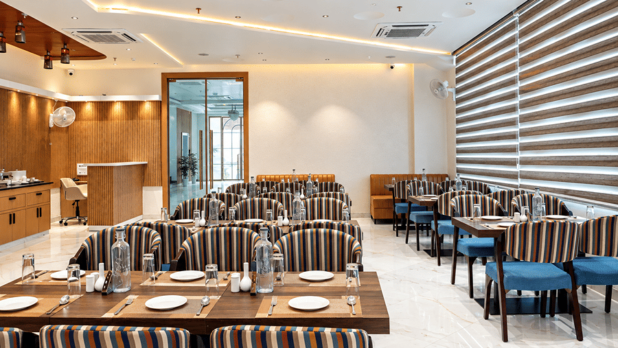 Restaurant seating area with wooden panelling, striped chairs, and window blinds at Daksh The Royal Braj, Vrindavan.