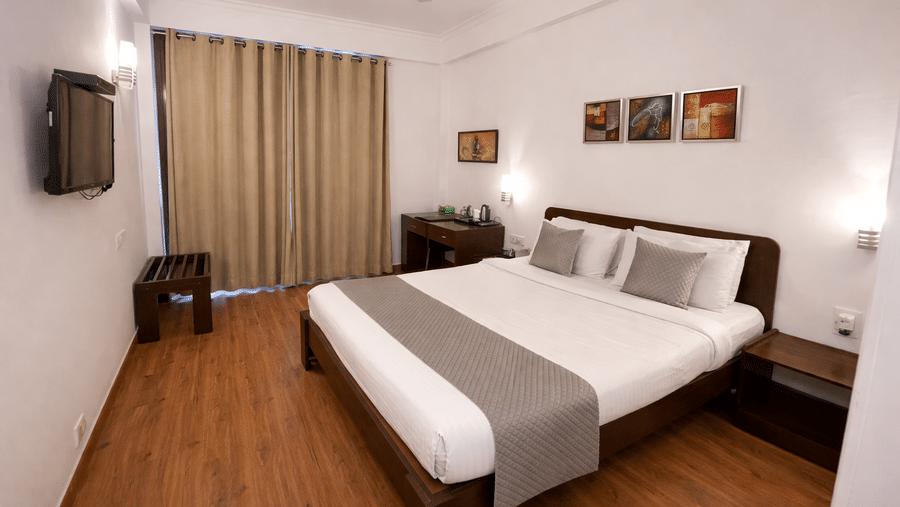 A bedroom with a king size bed, wall-mounted television, ceiling fan, and a large window at EMBLEM - Arjun Marg, Golf Course Road, Gurgaon.