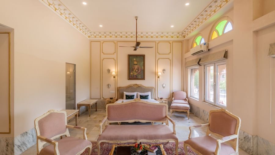 Front view of Maharaja Suite at Umaid Palace featuring a comfortable sitting area with sofa set and centre table. There is a bed behind the sofa with lamps on wither side, and two windows.