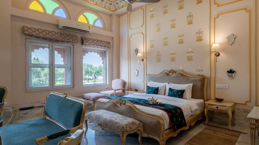 Maharaja Suite at Umaid Palace featuring a lavish bedroom with king size bedside table and lamps on either side. The room also have few soft sofa chairs an, a table, and a window near AC.
