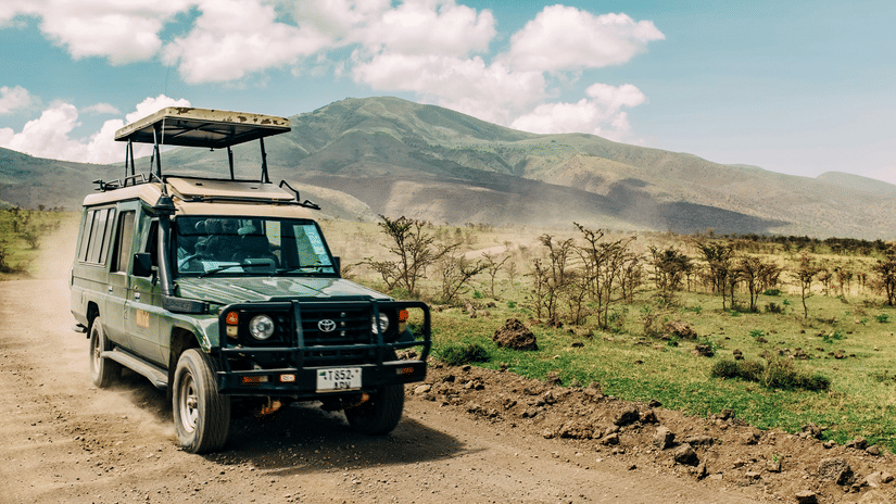 A safari jeep cruises through a dirt track, surrounded by open grasslands and distant rolling hills.