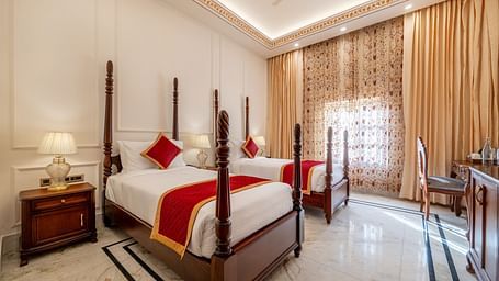 corner view image of a twin bed room along with a work desk and chair with tall golden curtains covering the window - Brij Castle, Jaipur